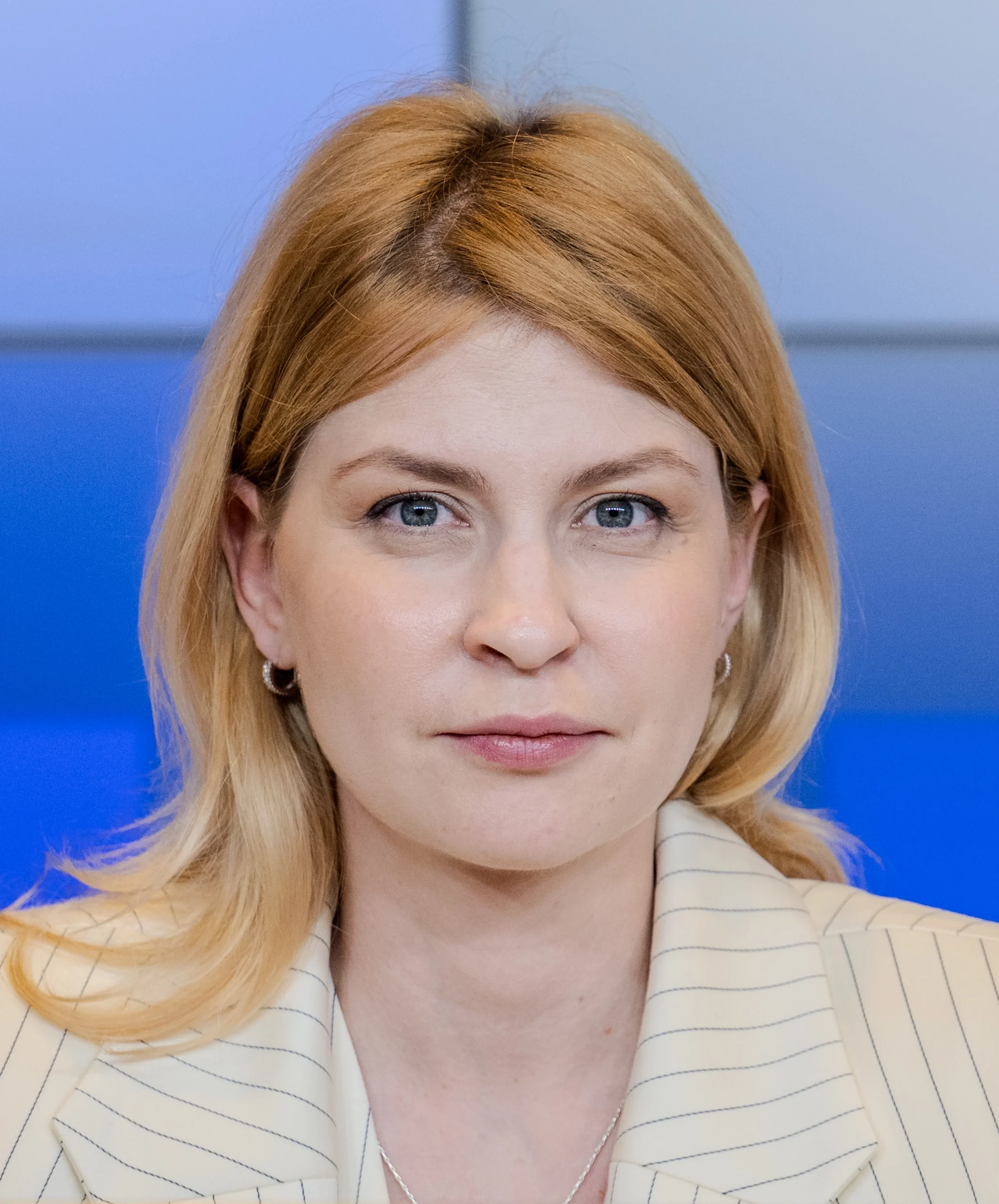Жінка з русявим волоссям у кремовому піджаку на синьому тлі