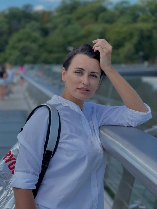 Жінка в білій сорочці з рюкзаком спирається на перила мосту на тлі зелені